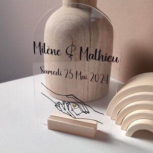 May include: A clear acrylic sign with a rounded top and a wooden base. The sign features a black illustration of two hands holding a heart. The text on the sign reads "Milène & Mathieu Samedi 25 Mai 2024".