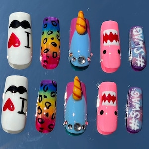 Könnte beinhalten: Ein Set aus zehn künstlichen Nägeln mit verschiedenen Designs. Die Designs umfassen einen weißen Nagel mit Schnurrbart und Herz, einen Regenbogen-Leopardenmuster, ein Einhorn, einen Hai und einen Nagel mit dem Text "#SWAG".