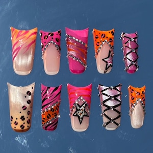Könnte beinhalten: Eine Sammlung von zehn künstlichen Fingernägeln mit verschiedenen Designs. Die Nägel zeigen eine Reihe von Farben, darunter Pink, Orange und Schwarz, mit Mustern wie Animal-Print, Streifen und Sternen. Einige Nägel sind mit Strasssteinen verziert.