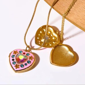 May include: A gold heart-shaped locket necklace with a pink enamel heart pendant. The pendant is decorated with blue and pink rhinestones and a larger iridescent rhinestone in the center. The locket opens to reveal a plain gold interior.