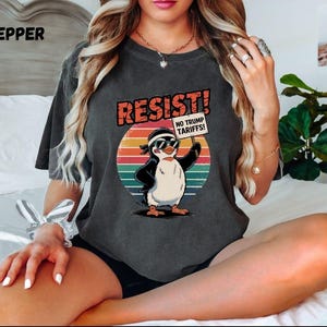 Puede incluir: Una camiseta gris oscuro con un gráfico de estilo vintage de un pingüino con gafas de sol y sosteniendo un cartel que dice "¡Resistir! ¡No a los aranceles de Trump!"