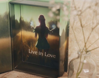 May include: A green box with a card inside. The card features a woman with wings standing in a field. The text on the card reads "Live in Love".