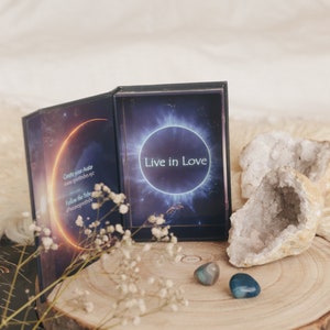 Puede incluir: Una caja cerrada de cartas de oráculo con un fondo azul oscuro y un círculo blanco con el texto "Live in Love" en el centro. La caja está abierta para revelar una carta con un diseño similar. La caja está sentada sobre una superficie de madera con dos pequeñas piedras azules y grises.