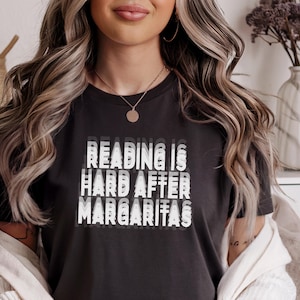 May include: Dark gray t-shirt with the text "READING IS HARD AFTER MARGARITAS" in a layered white font. The shirt is a crew neck style.