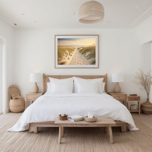 May include: A white bedroom with a large window overlooking a beach. The bed has white linens and a wooden headboard. There is a framed photograph of a beach scene above the bed. There are two nightstands with lamps on either side of the bed. A wooden bench sits in front of the bed.