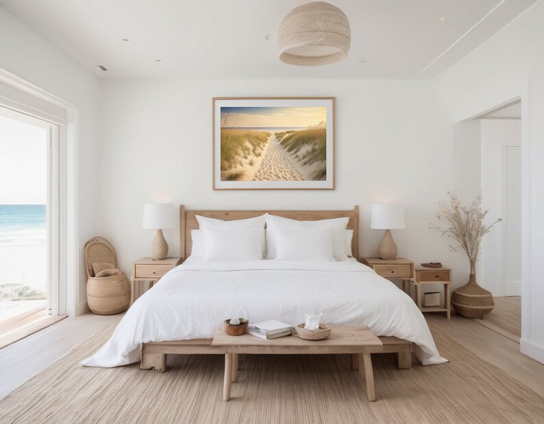 May include: A white bedroom with a large window overlooking a beach. The bed has white linens and a wooden headboard. There is a framed photograph of a beach scene above the bed. There are two nightstands with lamps on either side of the bed. A wooden bench sits in front of the bed.