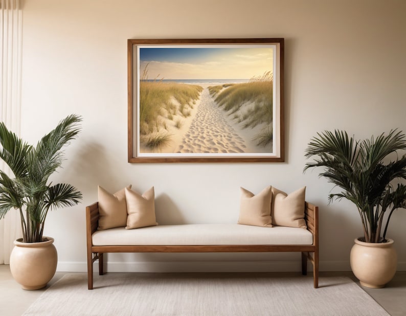 May include: A light wood bench with two beige cushions sits in front of a framed photograph of a sandy path leading to the ocean. Two potted palm trees flank the bench.