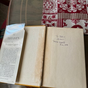 May include: An open book titled "Breakfast at Tiffany's" by Truman Capote, with a handwritten inscription dated June 1959. The book's cover is visible, along with a dust jacket. The book is on a table with a patterned tablecloth.