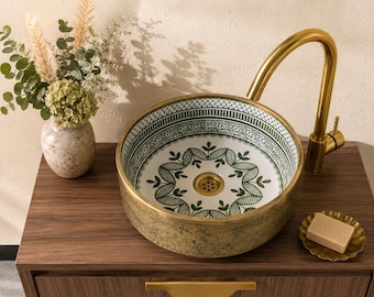 May include: A round vessel sink with a white interior featuring a green botanical pattern and a textured gold exterior. The sink includes a gold drain and a tall, curved gold faucet. A small bar of soap sits on a gold dish next to the sink.