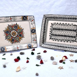 Peut inclure: Deux plats de service rectangulaires en céramique avec des motifs peints à la main. L'un présente un motif floral central en orange, vert et noir. L'autre a un motif géométrique noir et blanc. Des éléments décoratifs entourent les plats.