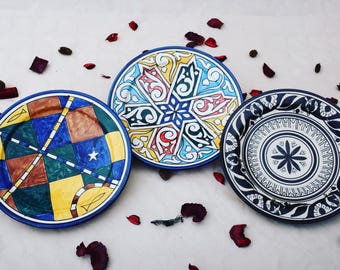 May include: Three ceramic plates with different colorful patterns. The first plate has a blue, yellow, green, and brown geometric pattern. The second plate has a blue, yellow, red, and black floral pattern. The third plate has a black and white floral pattern.