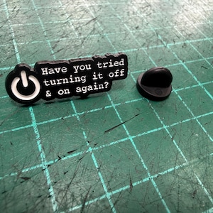 Może przedstawiać: Czarno-biała emaliowana przypinka z napisem "Have you tried turning it off & on again?" i symbolem przycisku zasilania. Przypinka znajduje się na zielonej powierzchni.