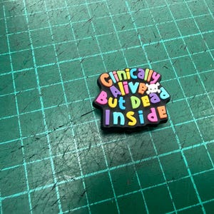 Pu&ograve; includere: Un charm per scarpe multicolore con la scritta "Clinically Alive But Dead Inside". Un piccolo teschio bianco &egrave; posto tra le parole "Alive" e "But". Il charm &egrave; su una superficie verde con un motivo a griglia.