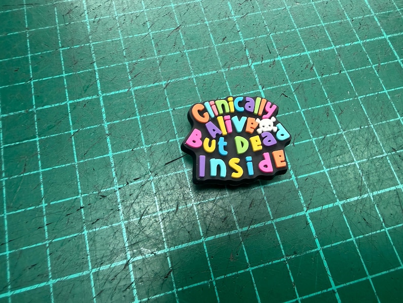 Pu&ograve; includere: Un charm per scarpe multicolore con la scritta "Clinically Alive But Dead Inside". Un piccolo teschio bianco &egrave; posto tra le parole "Alive" e "But". Il charm &egrave; su una superficie verde con un motivo a griglia.