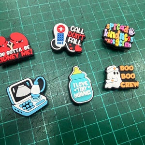Può includere: Una collezione di ciondoli per scarpe a tema medico. I ciondoli presentano vari disegni, tra cui un paio di reni con la scritta "YOU GOTTA BE KIDNEY ME!", un telefono con "CALL DON'T FALL" e un fantasma con "BOO BOO CREW".