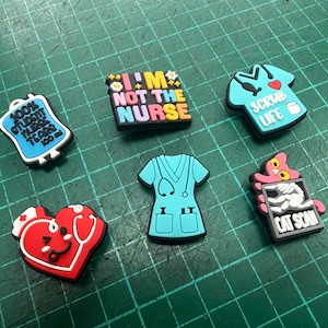 Può includere: Una collezione di ciondoli colorati a tema medico. I disegni includono un cuore con uno stetoscopio, un'uniforme da infermiera, una sacca di sangue e il testo che dice "I'M NOT THE NURSE", "SCRUB LIFE" e "CAT SCAN".