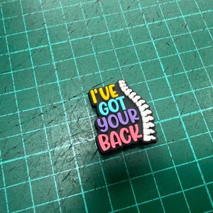 Pu&ograve; includere: Un ciondolo colorato per scarpe con la frase "I've Got Your Back" in giallo, blu, viola e rosa. Una grafica della colonna vertebrale bianca &egrave; sul lato destro del ciondolo. Il ciondolo &egrave; su una superficie verde.