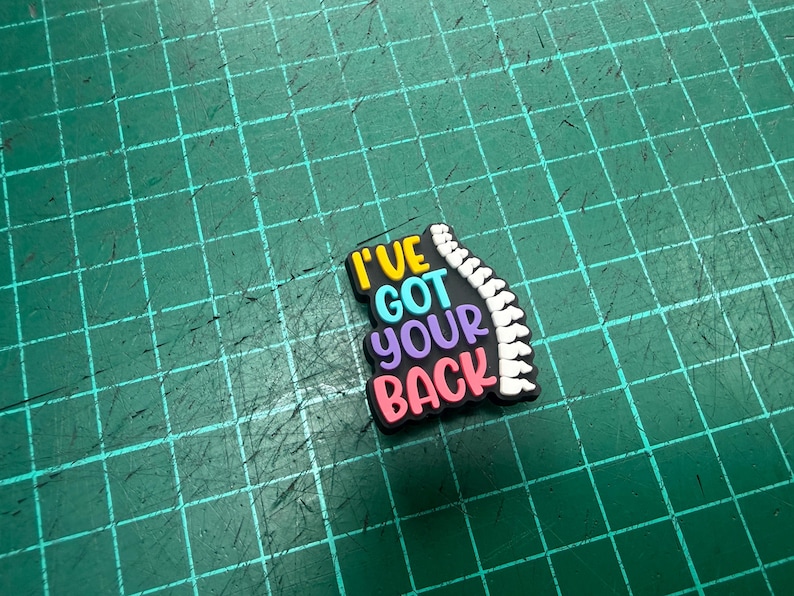 Pu&ograve; includere: Un ciondolo colorato per scarpe con la frase "I've Got Your Back" in giallo, blu, viola e rosa. Una grafica della colonna vertebrale bianca &egrave; sul lato destro del ciondolo. Il ciondolo &egrave; su una superficie verde.