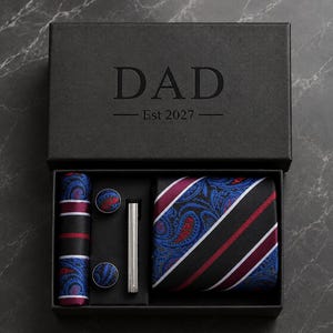 Può includere: Una scatola regalo nera con la scritta DAD Est 2027 contenente una cravatta piegata, un fazzoletto da taschino, due gemelli rotondi e un fermacravatta color argento. La cravatta e il fazzoletto presentano un motivo a righe e paisley blu, rosso e nero.