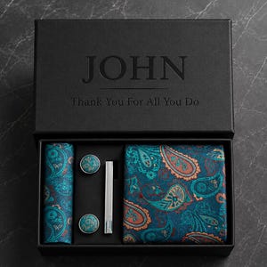 Può includere: Una scatola regalo nera con il testo JOHN e Thank You For All You Do, contenente una pochette, una cravatta, due gemelli rotondi con motivo paisley blu ottanio e arancione e un fermacravatta color argento.