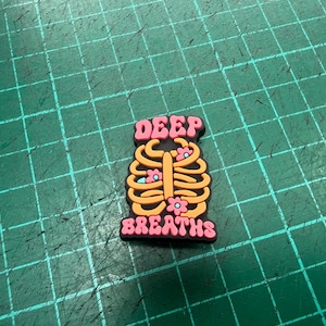 Pu&ograve; includere: Un ciondolo per scarpe con un design a gabbia toracica arancione e gialla, con piccoli fiori rosa. Le parole "DEEP" e "BREATHS" sono in rosa, con un contorno nero.