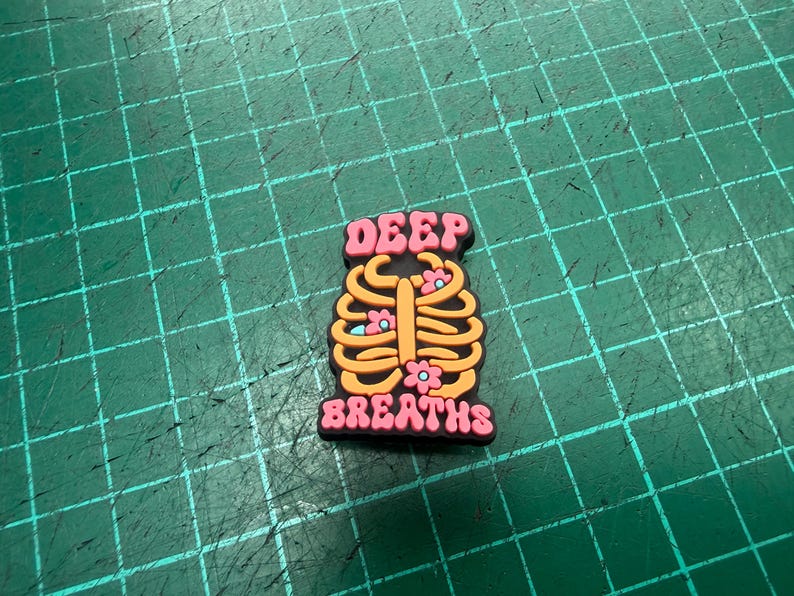 Pu&ograve; includere: Un ciondolo per scarpe con un design a gabbia toracica arancione e gialla, con piccoli fiori rosa. Le parole "DEEP" e "BREATHS" sono in rosa, con un contorno nero.