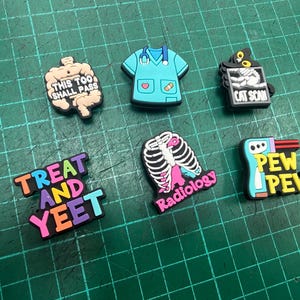 Può includere: Una collezione di ciondoli colorati a tema medico. Include un ciondolo con camice medico azzurro chiaro, un ciondolo nero con un'immagine di scansione TC e un ciondolo con la scritta "Treat and Yeet".