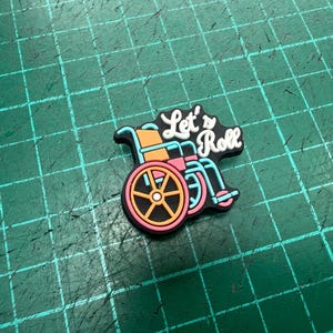 Pu&ograve; includere: Un ciondolo colorato con un design a sedia a rotelle con le parole "Let's Roll". La sedia a rotelle &egrave; raffigurata in arancione, rosa e blu, con un contorno nero. Il testo &egrave; in bianco e nero.