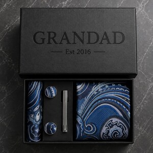 Può includere: Una scatola regalo nera con la scritta GRANDAD Est 2016 stampata sul coperchio. All'interno si trovano una cravatta con motivo paisley blu e grigio, una pochette abbinata, due gemelli rotondi con lo stesso motivo e un fermacravatta in metallo argentato.