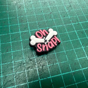 Pu&ograve; includere: Un ciondolo per scarpe con la frase "Oh Snap!" in rosa e bianco, con un design di ossa rotte. Il ciondolo ha un bordo nero, su uno sfondo a griglia verde.