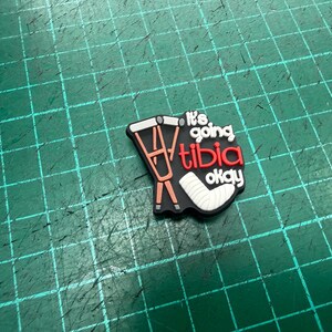 Pu&ograve; includere: Un ciondolo per scarpe nero con la scritta "It's going tibia okay" in bianco e rosso. Il ciondolo presenta un'immagine a cartoni animati di stampelle e un gesso per la gamba. Lo sfondo &egrave; una griglia verde.
