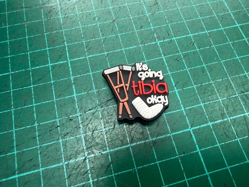 Pu&ograve; includere: Un ciondolo per scarpe nero con la scritta "It's going tibia okay" in bianco e rosso. Il ciondolo presenta un'immagine a cartoni animati di stampelle e un gesso per la gamba. Lo sfondo &egrave; una griglia verde.