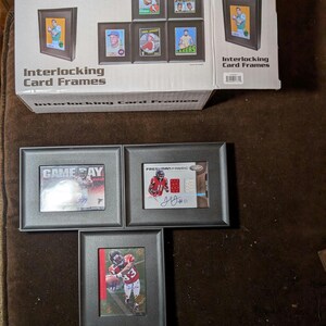 May include: A set of black interlocking card frames with a white box that says "Interlocking Card Frames". The frames are displaying various sports cards, including a baseball card with the text "LAKERS" and a football card with the text "FRESHMAN FABRIC".