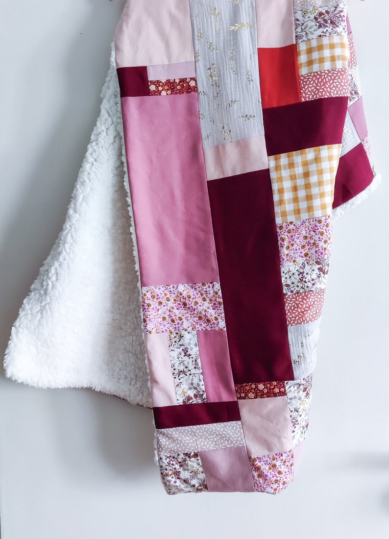 Może przedstawiać: Kołdra patchworkowa z r&oacute;żnymi wzorami kwiatowymi i geometrycznymi w odcieniach r&oacute;żu, bordowego i złotego. Kołdra jest wykonana z miękkich tkanin i idealnie nadaje się do przytulania na kanapie.