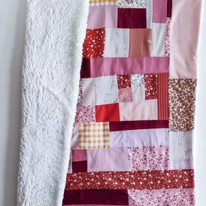 Może przedstawiać: Kołdra patchworkowa z r&oacute;żnymi wzorami kwiatowymi i geometrycznymi w odcieniach r&oacute;żowego, czerwonego i brązowego. Kołdra jest wykonana z r&oacute;żnych tkanin i faktur, w tym miękkiego, białego polaru.