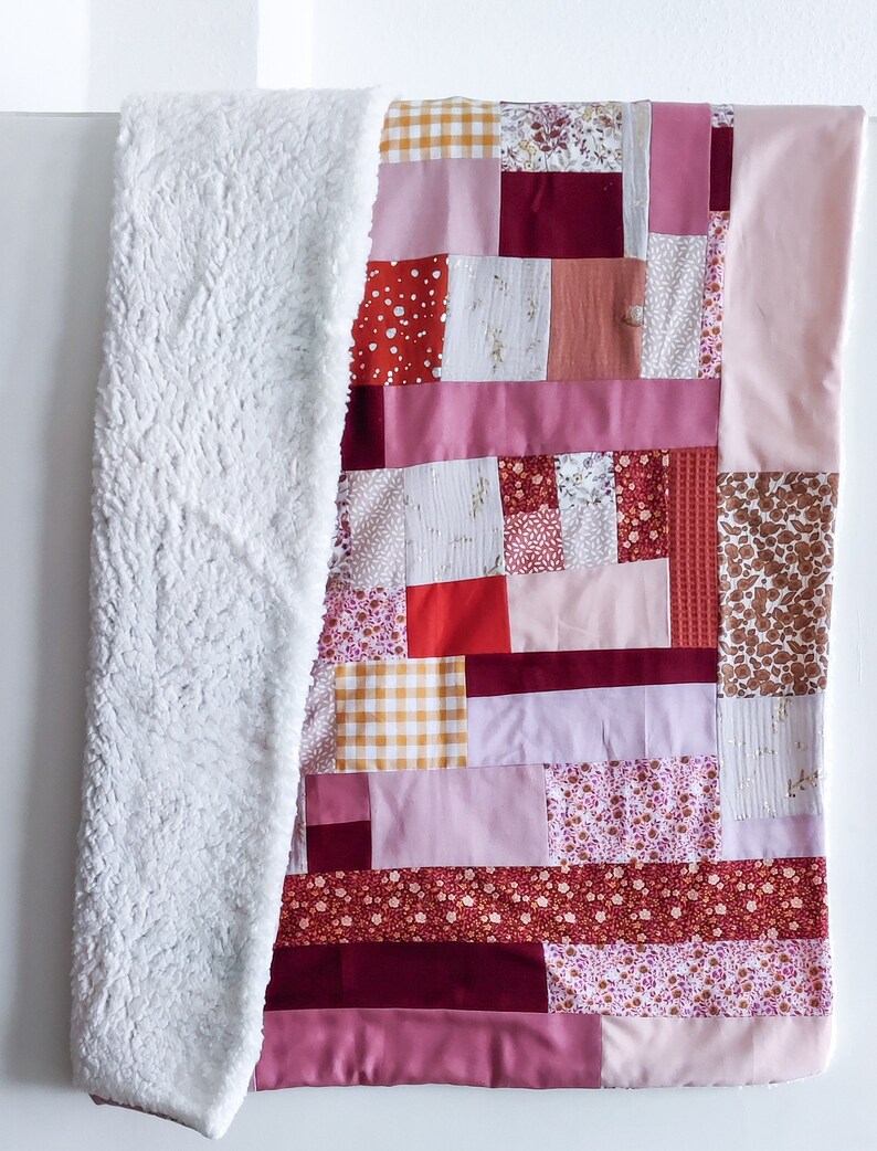 Może przedstawiać: Kołdra patchworkowa z r&oacute;żnymi wzorami kwiatowymi i geometrycznymi w odcieniach r&oacute;żowego, czerwonego i brązowego. Kołdra jest wykonana z r&oacute;żnych tkanin i faktur, w tym miękkiego, białego polaru.