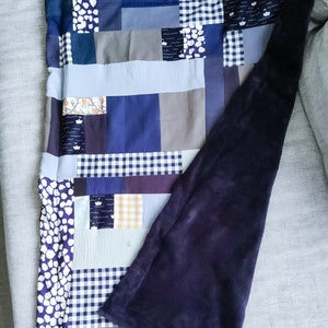 Może przedstawiać: Kołdra patchworkowa z granatowym podszewka. Kołdra wykonana jest z r&oacute;żnych tkanin w odcieniach niebieskiego, szarego i białego. Kołdra jest złożona na p&oacute;ł i leży na szarej kanapie.