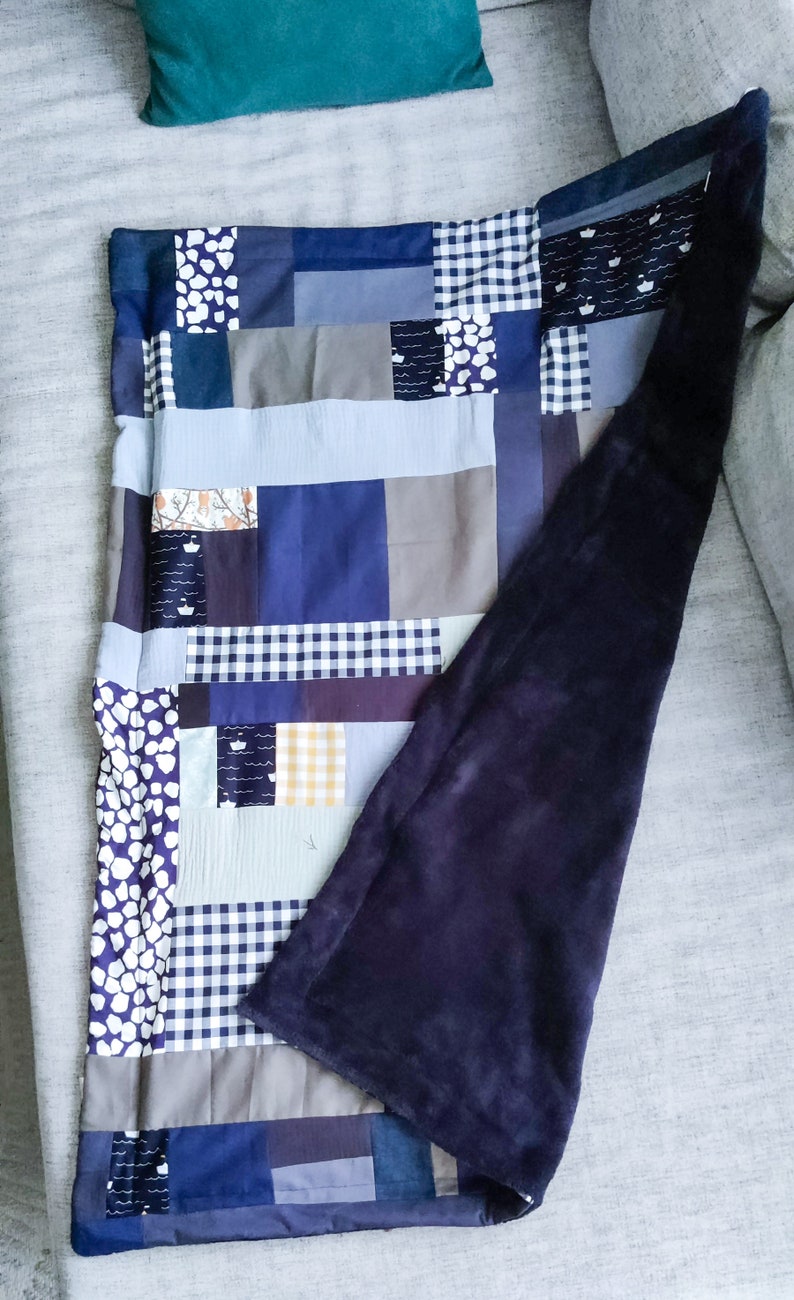 Może przedstawiać: Kołdra patchworkowa z granatowym podszewka. Kołdra wykonana jest z r&oacute;żnych tkanin w odcieniach niebieskiego, szarego i białego. Kołdra jest złożona na p&oacute;ł i leży na szarej kanapie.