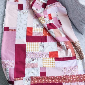 Może przedstawiać: Kołdra patchworkowa z r&oacute;żnymi wzorami kwiatowymi i geometrycznymi w odcieniach r&oacute;żowego, czerwonego i białego. Kołdra jest wykonana z r&oacute;żnych tkanin i faktur, tworząc unikalny i interesujący wz&oacute;r.