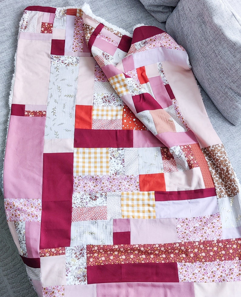 Może przedstawiać: Kołdra patchworkowa z r&oacute;żnymi wzorami kwiatowymi i geometrycznymi w odcieniach r&oacute;żowego, czerwonego i białego. Kołdra jest wykonana z r&oacute;żnych tkanin i faktur, tworząc unikalny i interesujący wz&oacute;r.