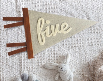 May include: A triangular felt pennant in light beige with the word five in cream-colored script, featuring a brown vertical strip with three horizontal tabs. A small crocheted white rabbit and a circular rattle with grey and green spheres.