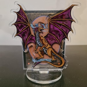 Puede incluir: Un soporte de acrílico transparente con un dragón con alas moradas y cuerpo marrón. El dragón está sobre un fondo con el texto "The Gathering". El soporte está diseñado para sostener una tarjeta.
