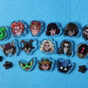 Peut inclure: Une collection de petites épinglettes en acrylique transparent représentant divers visages de personnages illustrés, une icône circulaire verte et un emblème noir en forme d'araignée. Des attaches en forme d'étoile noire sont éparpillées.