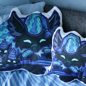 May include: Two plush pillows with a whimsical design. The pillows feature a dark blue creature with large ears and a patterned hat. The creature is surrounded by smaller versions of itself, all in shades of blue and teal.