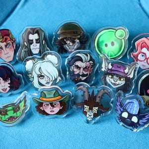 Puede incluir: Colección de pines acrílicos transparentes con retratos de personajes de dibujos animados. Los pines muestran una variedad de caras con diferentes expresiones, colores de pelo y accesorios. El fondo es de tela azul lisa.