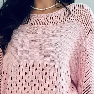 May include: A pink crochet sweater with a loose fit and a delicate open-stitch pattern. The sweater has a round neckline and three-quarter sleeves.