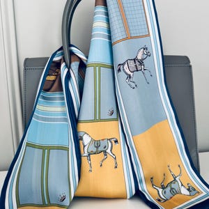 May include: A patterned silk scarf featuring horse illustrations. The scarf has a design with blue, yellow, and white panels, and is draped over a grey handbag. The scarf's design includes images of horses.
