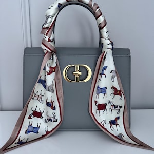 Op de afbeelding: Een grijze handtas met een goudkleurige sluiting en een handvat van een sjaal met patroon. De sjaal heeft een witte achtergrond met kleurrijke paardenillustraties en een roze rand. De tas is rechthoekig en heeft een gestructureerd ontwerp.