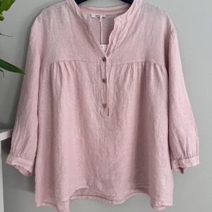 Pode incluir: Uma blusa de linho rosa claro com gola mandarim e três botões. As mangas três quartos e o corte folgado criam um estilo casual e confortável. A blusa está pendurada num cabide preto.