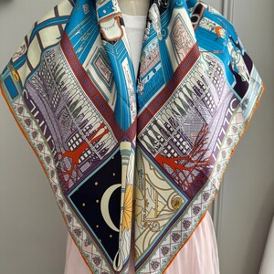 May include: A square silk scarf with a detailed architectural and celestial design. The scarf features a vibrant colour palette of blues, purples, and oranges, with an orange border. The design includes buildings, a crescent moon, and stars.